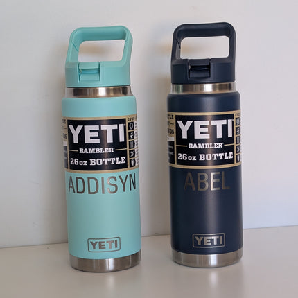 Two YETI water bottles, one light blue and one navy, engraved with names on a white surface with a white background.
