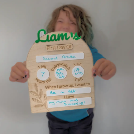 A child holding a personalized wooden First Day of School sign with the name 'Liam's' in green script, showing handwritten details for second grade and aspirations.