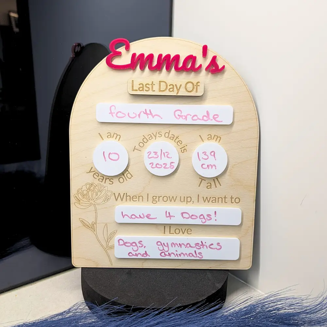 Personalized wooden milestone plaque for a child's last day of fourth grade, featuring editable white sticky note pads with handwritten details in pink and brown ink, displayed on a black stand.