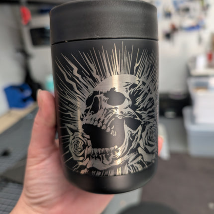 Black cooler with skull design held in a hand, blurred workshop background