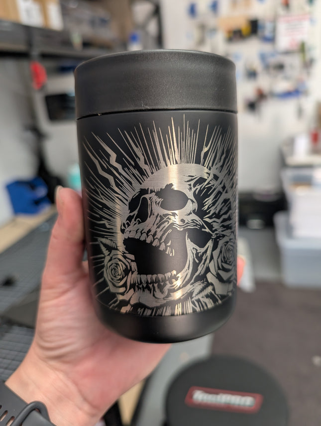 Black cooler with skull design held in a hand, blurred workshop background