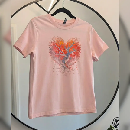 A light pink short-sleeve t-shirt hanging on a black hanger, featuring a graphic of a tree with orange, red, and blue leaves forming a heart shape on the chest. The shirt is displayed against a light gray wall with a circular black metal frame.