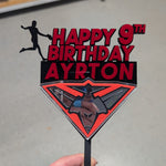 A hand holds a custom laser-cut cake topper featuring the text 'HAPPY 9TH BIRTHDAY AYRTON' in red and black, alongside a black silhouette of a soccer player and a red and black spider-themed diamond graphic at the bottom.