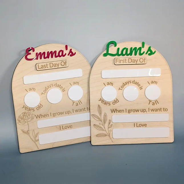 Two personalized wooden first-day-of-life milestone plaques on a gray background. One is labeled 'Emma's Last Day Of' with red lettering, and the other is labeled 'Liam's First Day Of' with green lettering.