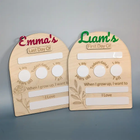 Two personalized wooden first-day-of-life milestone plaques on a gray background. One is labeled 'Emma's Last Day Of' with red lettering, and the other is labeled 'Liam's First Day Of' with green lettering.