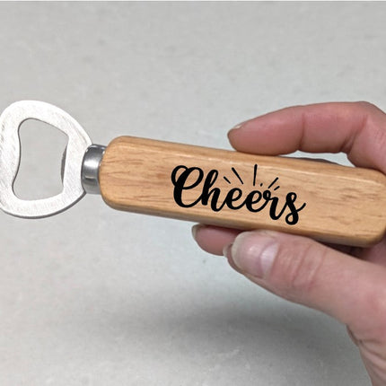 A hand holding a bottle opener with a light wood handle engraved with the word 'Cheers' in black script, set against a neutral gray background.