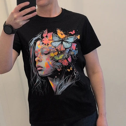 A person taking a mirror selfie wearing a black short-sleeve t-shirt with a colorful graphic print of a woman's face and butterflies.