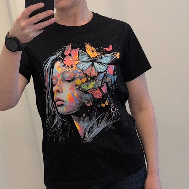 A person taking a mirror selfie wearing a black short-sleeve t-shirt with a colorful graphic print of a woman's face and butterflies.