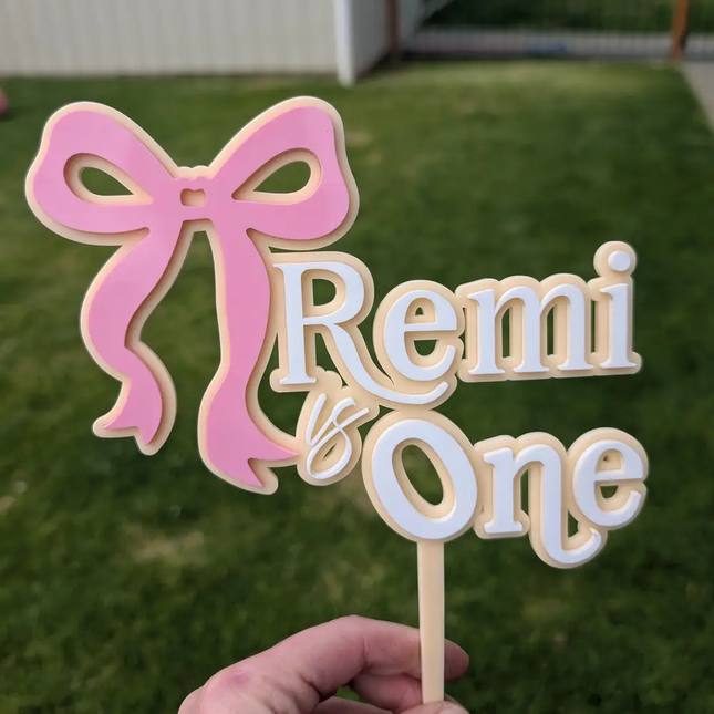 acrylic birthday cake topper with a pink bow and the text 'Remi is One' in white letters, held by a hand outdoors on a grassy lawn.