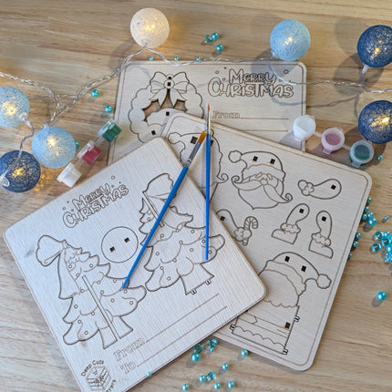 All 3 paint your own christmas boards arranged on a workbench with 2 paintbrushes and 3 paint pots with red, green and white paint in them.  there are blue christmas lights nd blue decorations around