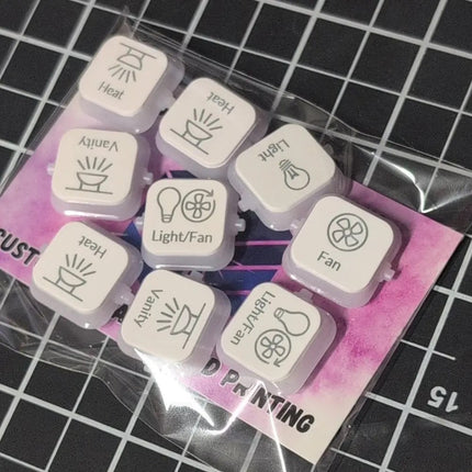 A set of nine small square white clipsal dollies with engraved icons and labels for Heat, Ventily, Light, Fan, and Light/Fan, packaged in a clear plastic sleeve over a pink backing card on a black grid surface.