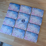 Twelve rclipsal iconic cover plates arranged in a grid on a light wood tabletop. Each case has a pastel watercolor-style floral design with pink blossoms and a blue-to-purple gradient background.
