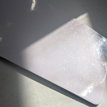 A close-up of a rectangular, slightly rounded-corner metallic surface with a pearlescent, glittery finish, resting on a light gray surface under direct sunlight.