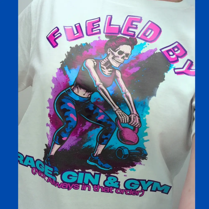 A close-up of a white graphic t-shirt featuring a colorful illustration of a skeleton exercising with a kettlebell. The graphic includes the text 'FUELED BY' in pink and 'RAGE: GIN & GYM (Not Always In that order)' in blue and pink.
