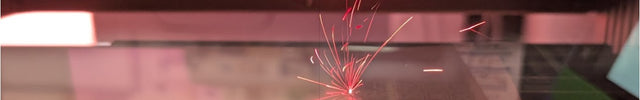 A close-up of a laser engraving or cutting process, showing bright red laser sparks and light streaks striking and marking a transparent surface, with a dark machine frame visible in the background.