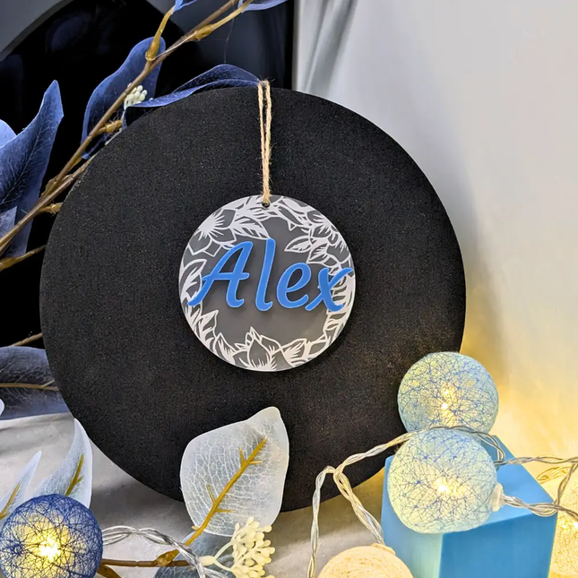 A personalized circular hanging ornament with a silver floral border and the name 'Alex' in blue script, displayed on a black round stand with decorative string lights and blue leaves around it.