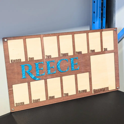 Wooden photo frame board with 'REECE' engraved in blue, against a gray wall designed to diplay school photos throughout all of school
