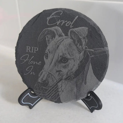 A round dark gray slate memorial plaque on a small black display stand, featuring an engraved portrait of a dog and the words 'Errol' and 'RIP Home In'.