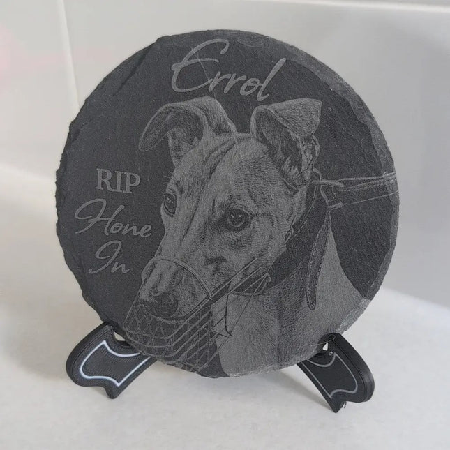 A round dark gray slate memorial plaque on a small black display stand, featuring an engraved portrait of a dog and the words 'Errol' and 'RIP Home In'.