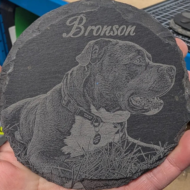 A round dark slate coaster engraved with a portrait of a pitbull-type dog and the name 'Bronson' in script, held in a person's hand.