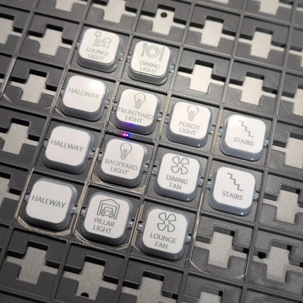 A close-up of a smart home security camera or smart lock keypad panel with multiple clipsal iconic dollies labeled for different functions like 'LOUNGE LIGHT', 'DINING LIGHT', 'HALLWAY', 'STAIRS', and 'LOUNGE FAN' on a dark textured grid background.