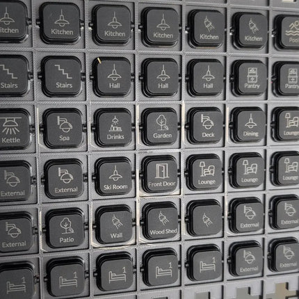 A close-up grid of extra black clipsal iconic dolly rockers category buttons with white icons and labels such as Kitchen, Stairs, Hall, Pantry, Kettle, Spa, Drinks, Garden, Deck, Dining, Lounge, External, Patio, Wall, Wood Shed, and Front Door.