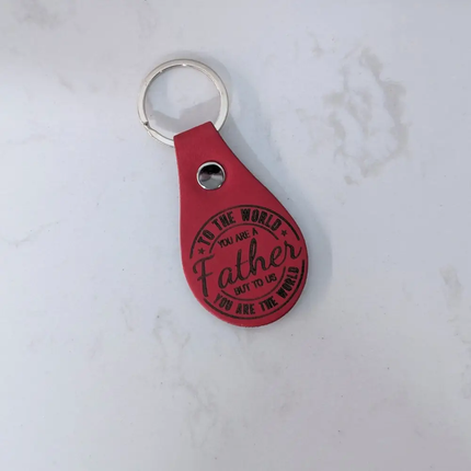 A red leather keychain with a silver metal key ring and a silver rivet, featuring the engraved text 'TO THE WORLD YOU ARE Father BUT TO US YOU ARE THE WORLD' in black lettering, set against a light marble background.