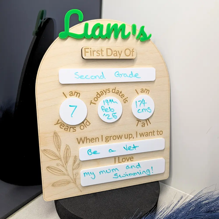 Personalized wooden First Day of School milestone plaque for Liam, featuring a green 'Liam's' script at the top, white milestone circles, and handwritten text celebrating second grade, displayed on a tabletop.