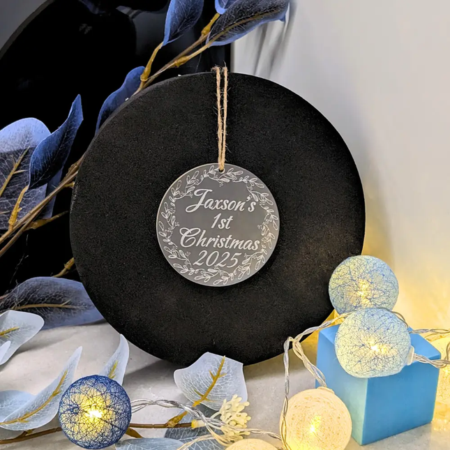 A black round decorative plaque or sign with a hanging twine loop and a silver circular ornament engraved with the personalized text 'Jaxson's 1st Christmas 2025' surrounded by a wreath design. The sign is styled with blue leaves, warm white string lights, and a small blue gift box in a holiday-themed setting.