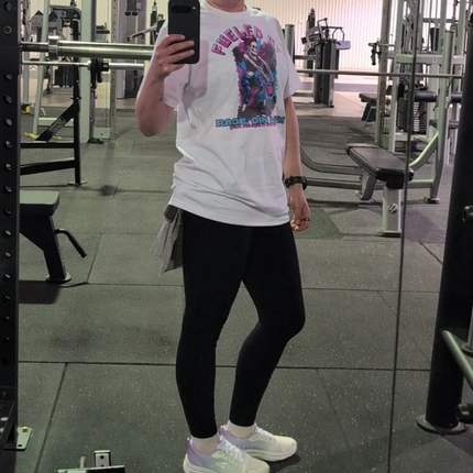 A person taking a mirror selfie in a gym, wearing a white graphic t-shirt with a colorful print, black leggings, and white sneakers.