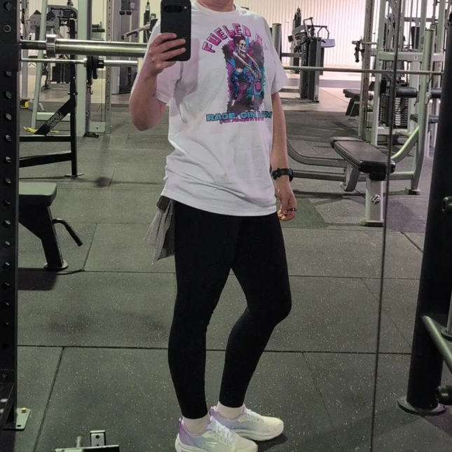 A person taking a mirror selfie in a gym, wearing a white graphic t-shirt with a colorful print, black leggings, and white sneakers.