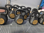 Several black kettlebells arranged on a gray gridded cutting mat, with round gold labels reading "Member Month" on the front of each kettlebell.