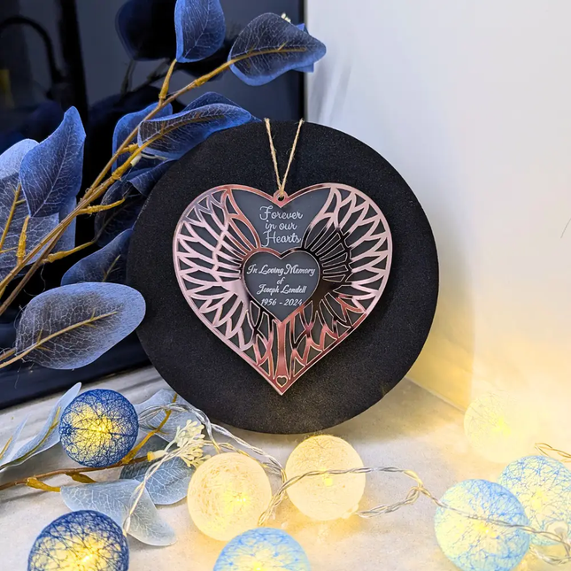 A heart-shaped hanging memorial ornament with engraved text, displayed on a round black stand with decorative blue leaves and warm string lights around it.