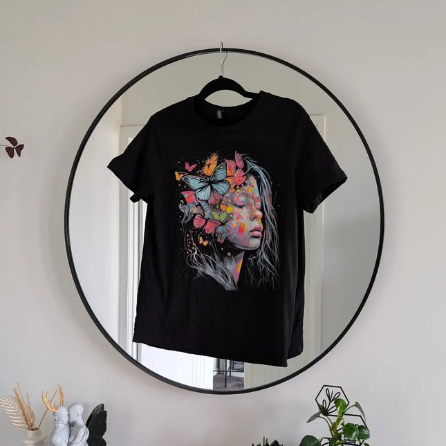 A black short-sleeve T-shirt hanging on a black hanger in front of a large round mirror. The shirt features a colorful graphic print of a woman's face with butterflies and flowers integrated into her hair.