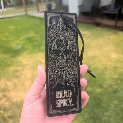 A hand holding a black leather bookmark or book tag with an embossed skull, roses, dagger, and the words 'READ SPICY.' The background shows a blurred outdoor lawn and patio area.