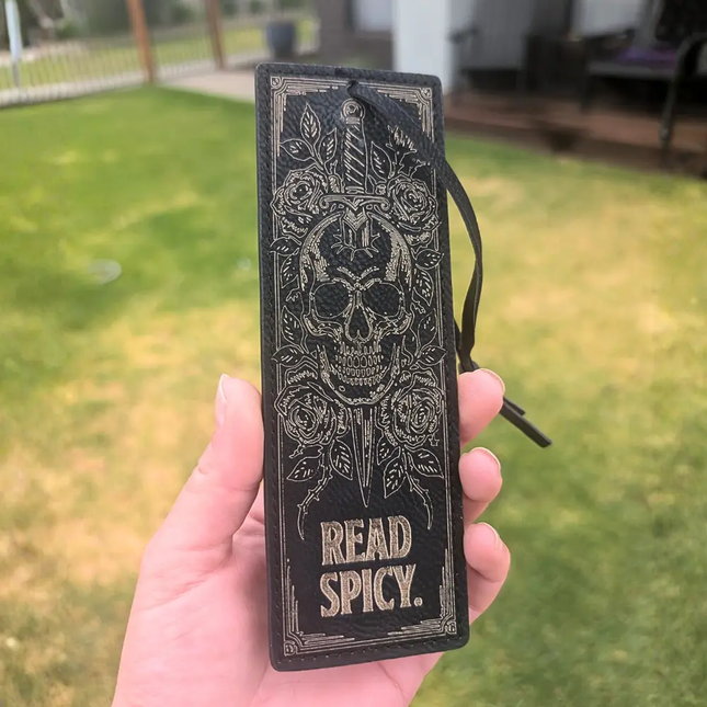 A hand holding a black leather bookmark or book tag with an embossed skull, roses, dagger, and the words 'READ SPICY.' The background shows a blurred outdoor lawn and patio area.