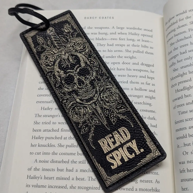 A black leather bookmark with silver-toned metal detailing, featuring a skull and roses design and the words 'READ SPICY.' at the bottom, resting on an open book.