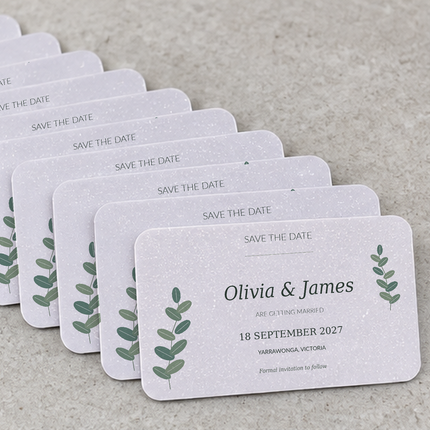 A set of white textured 'Save the Date' cards arranged in a fan shape on a neutral gray surface. Each card features a delicate green leafy sprig design on the left and right edges, with placeholder names and wedding details printed on the front.