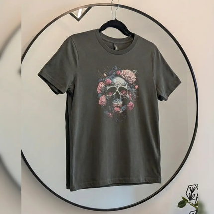 A dark gray crewneck t-shirt hanging on a black hanger inside a large circular mirror. The shirt features a graphic print of a skull surrounded by pink and red roses.