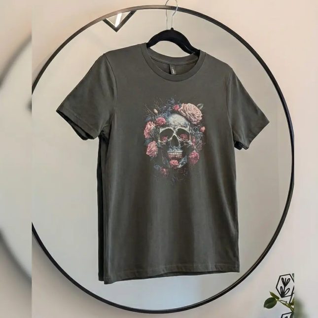 A dark gray crewneck t-shirt hanging on a black hanger inside a large circular mirror. The shirt features a graphic print of a skull surrounded by pink and red roses.