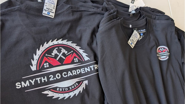 A stack of dark charcoal gray t-shirts with the Smyth 2.0 Carpenter logo printed on the front, laid out on a light-colored surface.