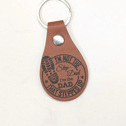 A brown leather keychain tag with a silver metal key ring and snap button, featuring a black printed design that reads 'I'M NOT THE Step-Dad I'm the DAD THAT STEPPED UP' with a footprint graphic, on a plain white background.