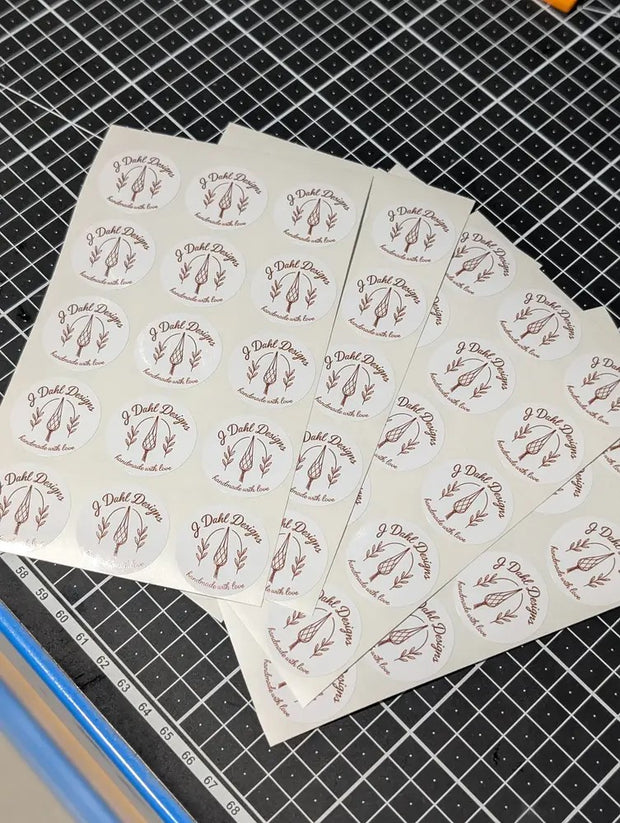 Several sheets of round white stickers printed with a repeating logo in dusty rose-brown, arranged on a black gridded cutting mat.