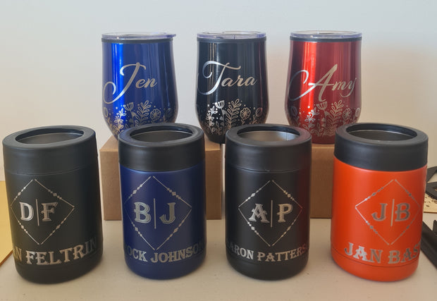 Six insulated tumblers with black lids are arranged vertically on a wooden display block against a plain light wall. The tumblers come in red, dark blue, and black finishes, each with custom printed names and monogram designs in gold or silver.