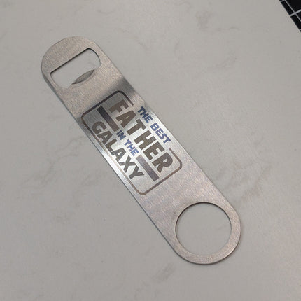 Metal bottle opener with 'Father of the Galaxy' text on a gray surface