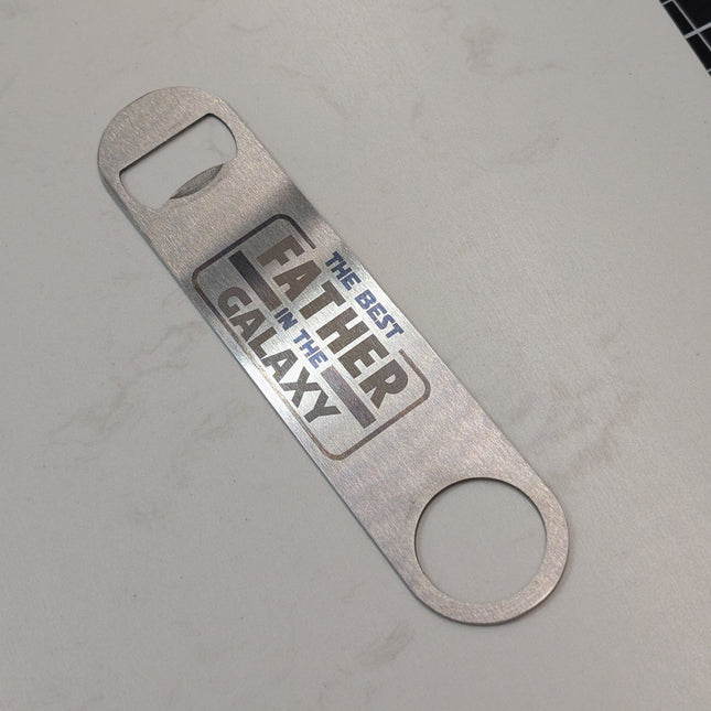 Metal bottle opener with 'Father of the Galaxy' text on a gray surface