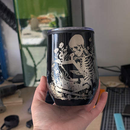 Hand holding a black tumbler with a white skeleton design against a blurred indoor background