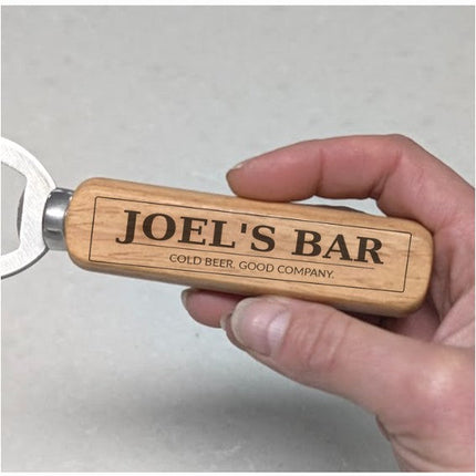 A hand holding a personalized wooden bottle opener with 'JOEL'S BAR' engraved on the handle, set against a neutral gray background.