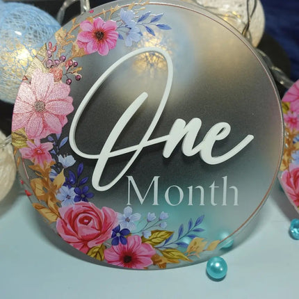 Decorative round sign with floral design and 'One Month' text on a light blue background made out frosted acrylic with UV printed decoration