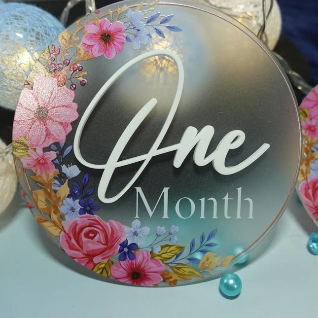 Decorative round sign with floral design and 'One Month' text on a light blue background made out frosted acrylic with UV printed decoration